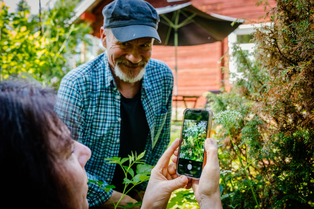 Kuvituskuvassa on kaksi henkilöä, joista toinen kuvaa kännykällä kasvia. Toinen henkilö katsoo kuvaajaa. Taustalla on punainen rakennus ja aurinkovarjo.