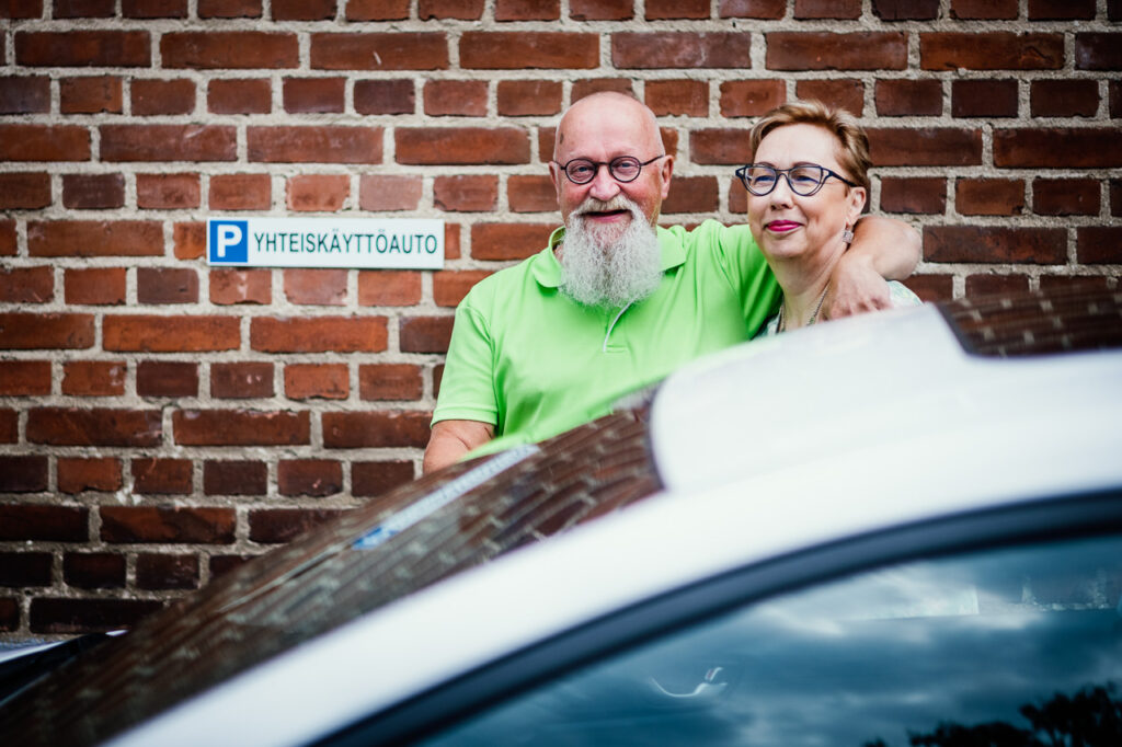 Kuvassa näkyy auton takana kaksi ihmistä. Seinässä on kyltti, jossa lukee P yhteiskäyttöauto.