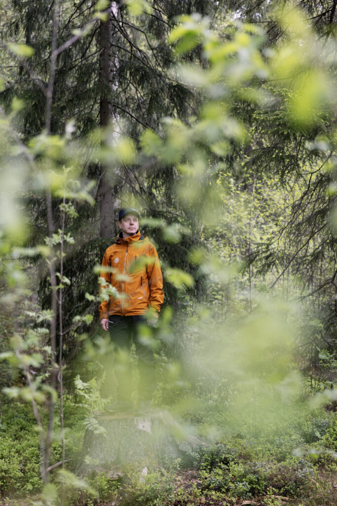 Lehvästön läpi näkyy oranssiin takkiin pukeutunut henkilö. Henkilö seisoo kannon päällä.