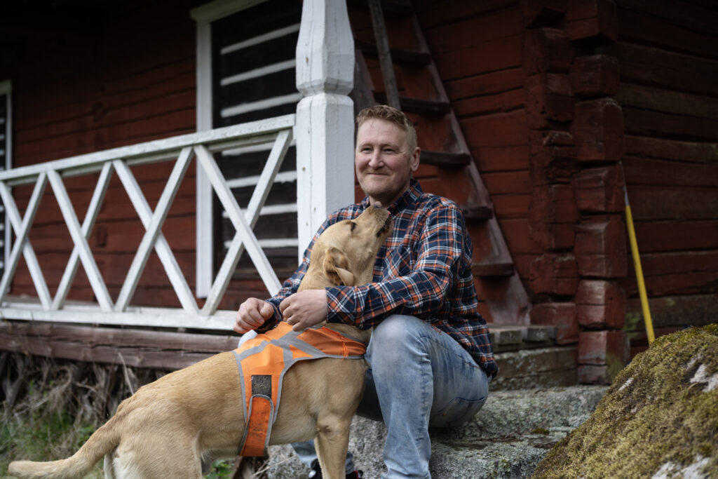 Ruutupaitainen mies istuu kivellä kädet koiran ympärillä. Taustalla punainen hirsirakennus. 