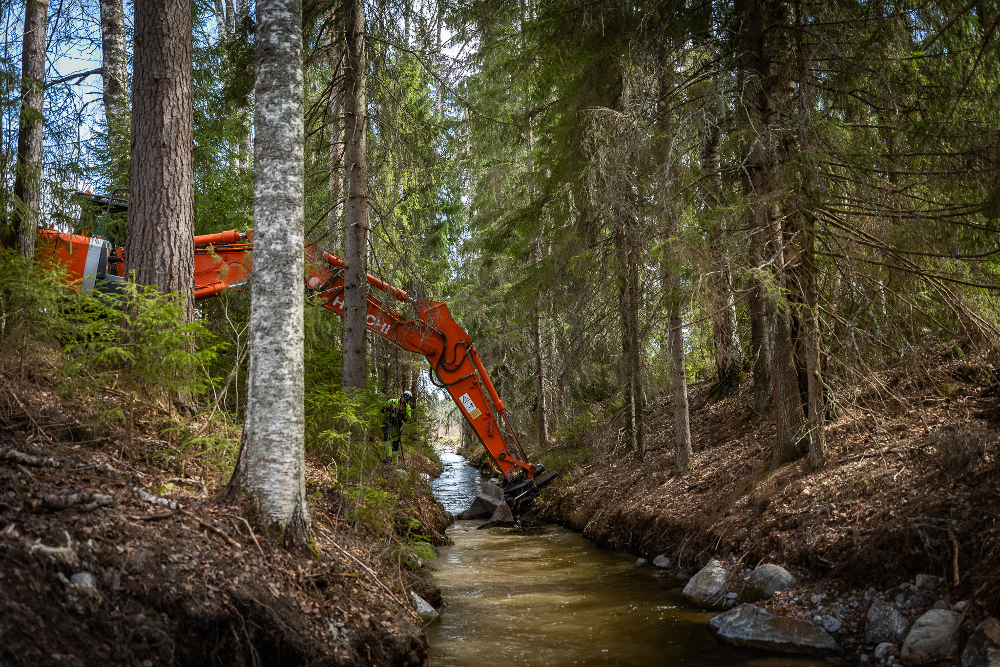Kaivinkone kaivaa puroa. Reunoilla puustoa.