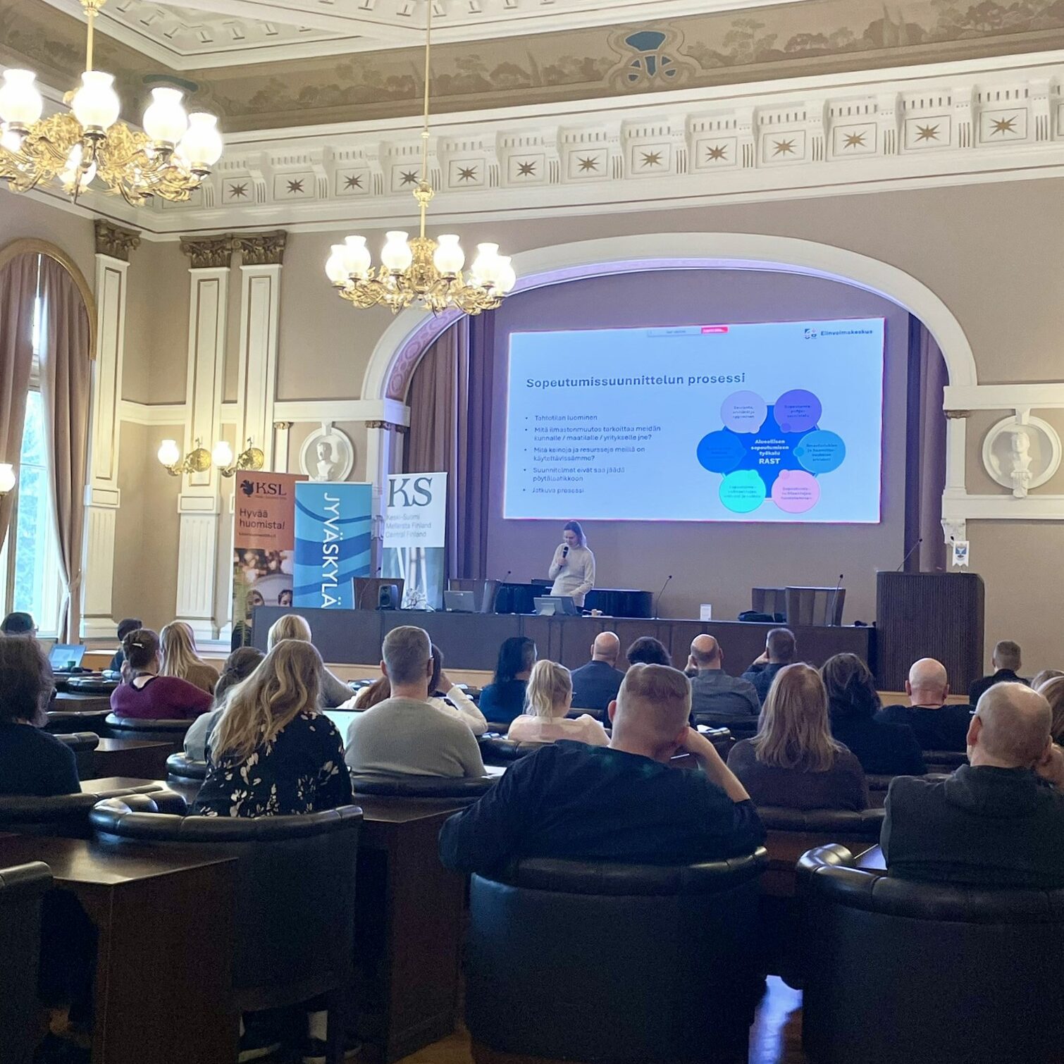 Koristeellisessa juhlasalissa järjestettävä seminaari. Yleisö istuu pöytien ääressä, ja salin edessä puhuja esittelee valkokankaalla sopeutumissuunnittelun prosessia.